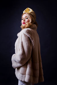 Portrait of beautiful woman wearing fur jacket and crown against black background