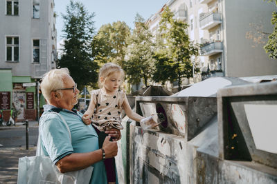 Senior man carrying granddaughter trashing garbage in can at street