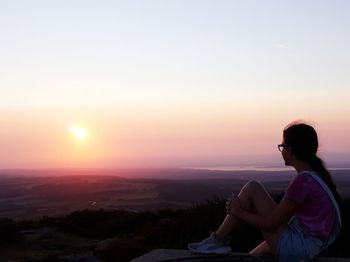 Side view of woman sitting on landscape against sky during sunset