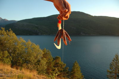 Cropped hand of person holding starfish with hook against lake and mountains