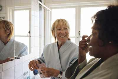 Happy blond woman talking to female friend while doing skincare in bathroom at home