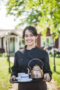 Portrait of confident young female waitress smiling while holding serving tray against restaurant