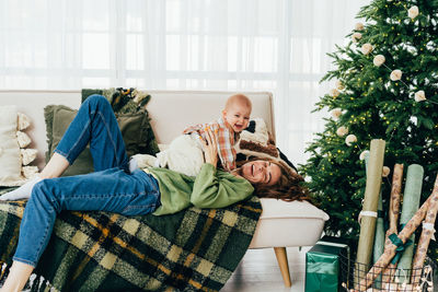 Happy mom and baby are lying on the couch next to the christmas tree and laughing