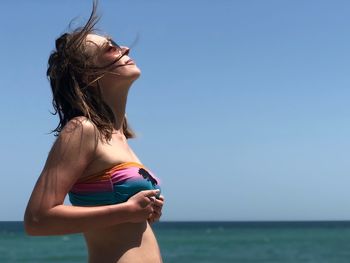 Woman in sunglasses and bikini standing by sea against clear sky