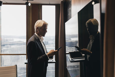 Side view of senior businessman using smart phone while standing by reflection on television in board room