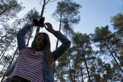 Low angle view of woman photographing through camera while standing against trees at forest