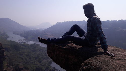 Man sitting on rock looking at mountains against sky