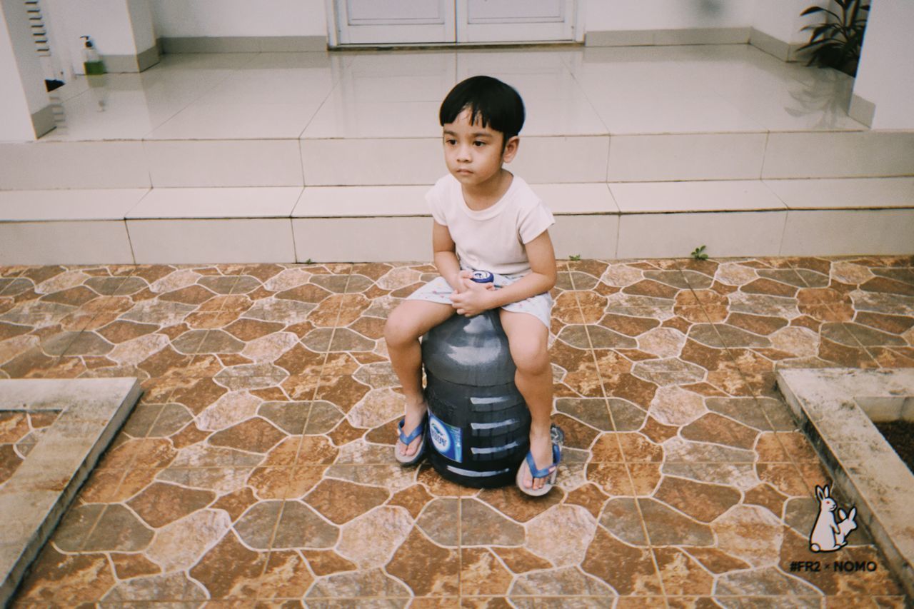 full length, childhood, one person, sitting, child, real people, front view, lifestyles, leisure activity, casual clothing, portrait, innocence, indoors, home interior, cute, males, looking at camera, flooring, tiled floor