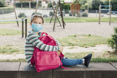 A girl in a protective mask sits in the schoolyard with her pink backpack. rest from study. 