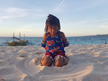 Portrait of girl kneeling on sand at beach against sky