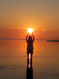 Silhouette woman with arms raised standing in sea against sky during sunset
