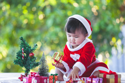 Full length of cute baby girl wearing santa costume while sitting with christmas decorations on table against trees