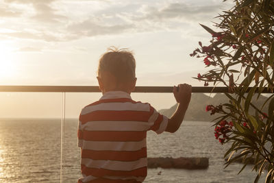 Rear view of boy standing by sea against sky during sunset