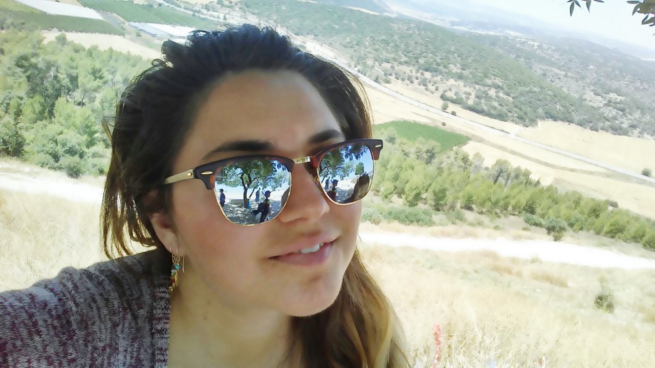 sunglasses, headshot, person, young adult, portrait, looking at camera, lifestyles, leisure activity, smiling, close-up, front view, sunlight, young women, focus on foreground, head and shoulders, day, field, sky