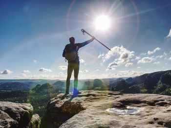 Tourist boy with backpack and sticks in mountain range standing on rocky top on blue morning sky