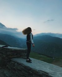 Side view of young woman standing on mountain against sky during sunset