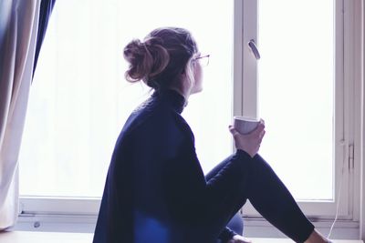 Side view of thoughtful woman having coffee while sitting on window sill