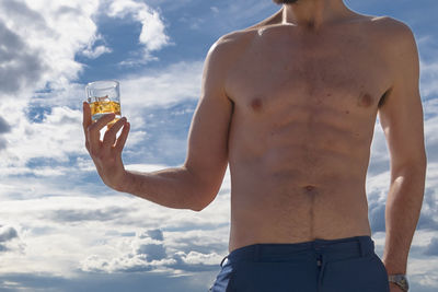 Midsection of shirtless man holding glass against sky