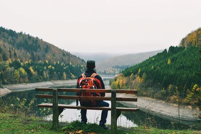 Rear view of man sitting on bench by lake against sky