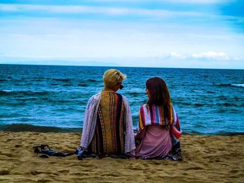 Rear view of women sitting on beach against sky