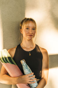 Portrait of woman with water bottle and exercise mat against wall