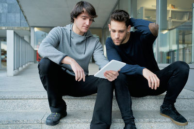 Front view of two young men talking sitting on a city staircase and lo