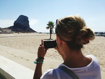 Rear view of woman photographing with mobile phone at beach against sky