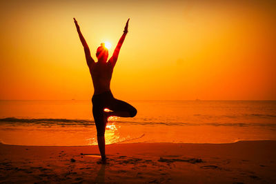 Silhouette woman with arms raised while doing yoga at beach against sky during sunset