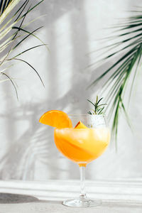 A glass with a summer cocktail with an orange and a sprig of rosemary under the branches of palm 