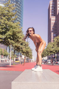 Young woman wearing bikini while laughing against buildings in city during sunny day