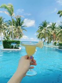 Cropped hand of woman holding drink in swimming pool against sky