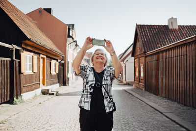 Smiling senior woman photographing while standing on road in city against clear sky