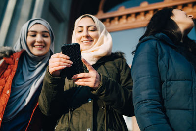 Low angle view of young female friends sharing smart phone by teenage girl in city