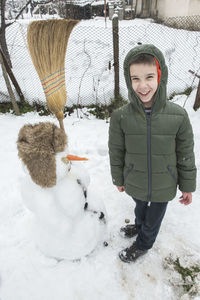 Full length of smiling boy standing by snowman in yard during winter