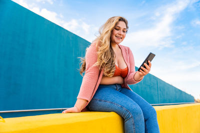 Young woman using mobile phone sitting on wall