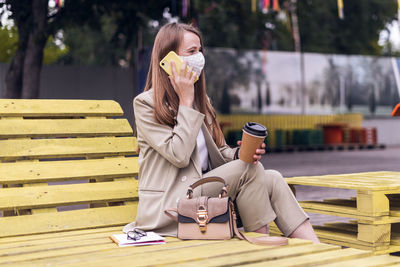 Beautiful business woman in medical face mask talking over the phone and sit down on bench outside.