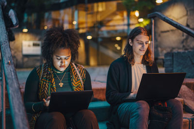 Young male and female students using laptop while sitting on steps in campus at night