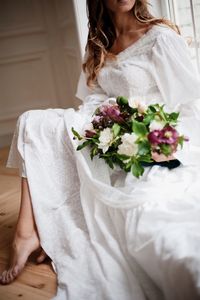 Low section of bride holding bouquet while sitting at window sill in room
