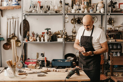 Serious middle aged male carpenter in apron messaging on mobile phone while standing near workbench with professional tools in creative work studio