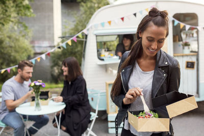 Female customer holding disposable salad box against food truck with friends and owner in background