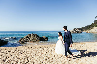 Newlywed couple walking at beach against clear sky during sunny day