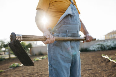 Senior man in denim holding shabby hoe tool and standing on cultivated land
