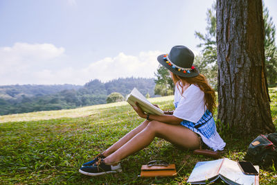 Woman with diary sitting by tree trunk on grassy land