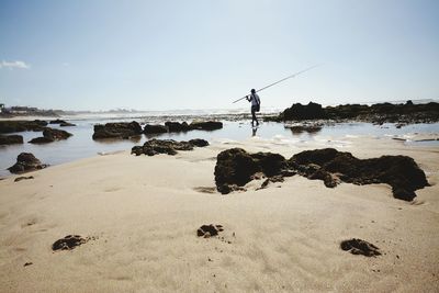 Rear view of man carrying fishing rod while walking on sea shore