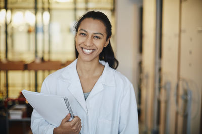 Portrait of smiling young chemistry student wearing lab coat standing with book in university