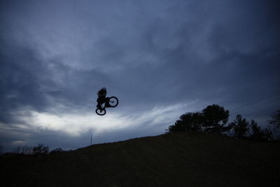 Low angle view of silhouette man performing bicycle stunt against cloudy sky