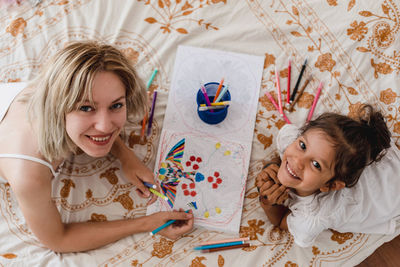 High view of caucasian mom and hispanic daughter painting while they are lying in the bed.