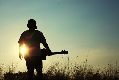 Silhouette man with guitar standing on field against sky during sunset