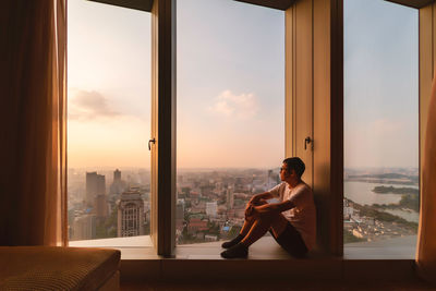 Man sitting on window sill during sunset