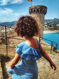 Rear view of girl with curly hair standing on steps against blue sky during sunny day